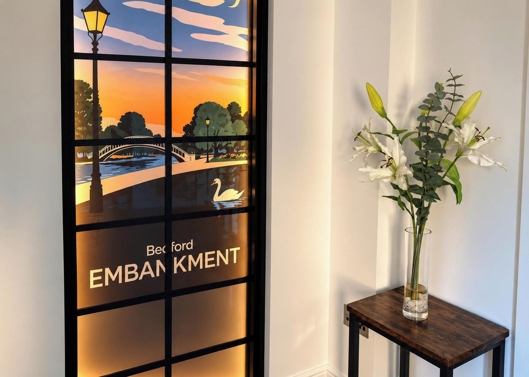A reception area with a decorative window featuring a sunset scene, a swan, and the text 'Bec ford EMBAN KMENT.' A vase with white flowers is placed on a wooden table beside the window.