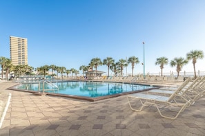 A swimming pool with lounge chairs and palm trees, surrounded by a paved area and a tall building in the background. - The Summit 1028 (Panama City Beach)