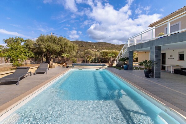A swimming pool with lounge chairs, a wooden fence, and a building with a tiled roof.
