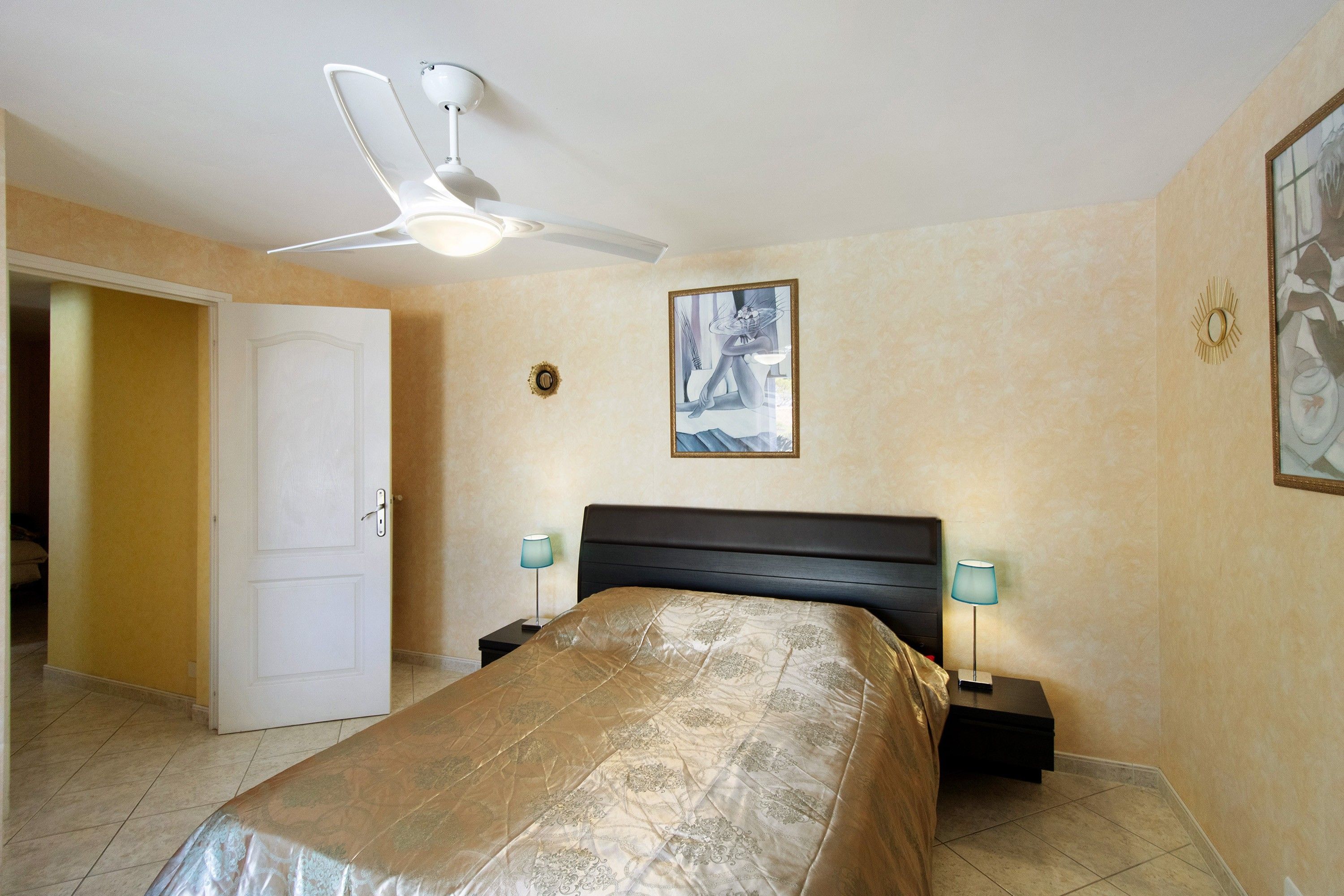 A bedroom with a bed, a ceiling fan, a painting, and a door.