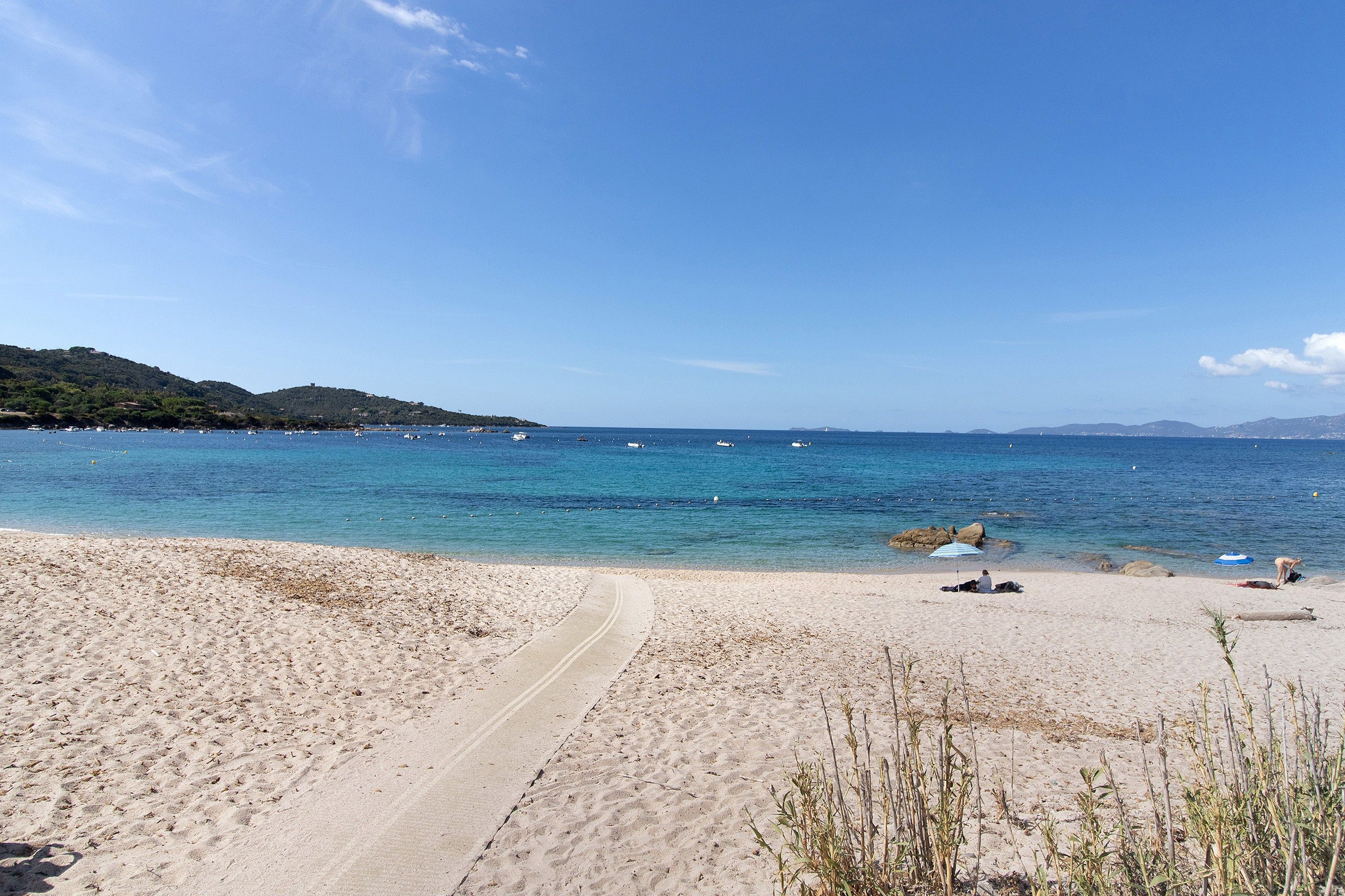 Une plage avec un chemin dégagé menant à l’eau, quelques personnes et des bateaux visibles au loin.