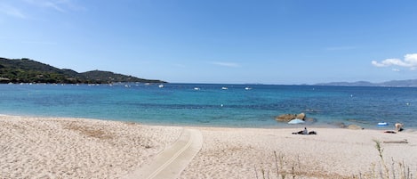 A beach with a clear path leading to the water, a few people, and boats in the distance.