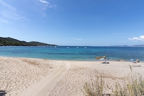 Une plage avec un chemin dégagé menant à l’eau, quelques personnes et des bateaux visibles au loin.
