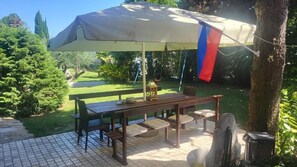 Une terrasse avec une table et des chaises, protégée par un grand parasol, entourée de verdure et d’une pelouse.
