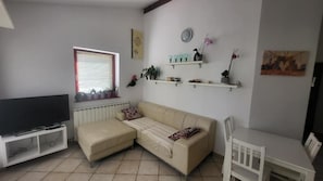 Un salon avec un canapé d’angle beige, un téléviseur posé sur un meuble, une table à manger avec des chaises et des étagères décoratives au mur.