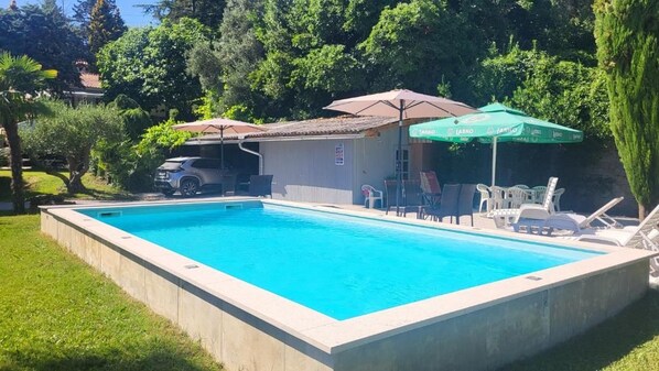 Une piscine rectangulaire, bordée d’une terrasse carrelée, entourée de pelouse et d’arbres. On y trouve des chaises longues et un parasol vert orné de logos.