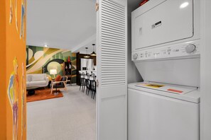 Eine moderne Küche mit Waschmaschine, einem Essbereich mit weißem Tisch und Stühlen sowie einem farbenfrohen, abstrakten Wandbild.