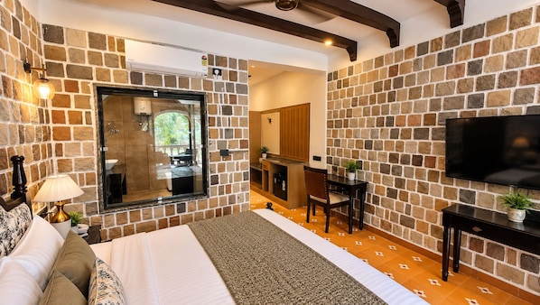 A hotel room with a stone wall, a bed, a desk, a television, and a view of the outside. - Luxuria Jim Corbett - By Moustache (Kaladhungi)
