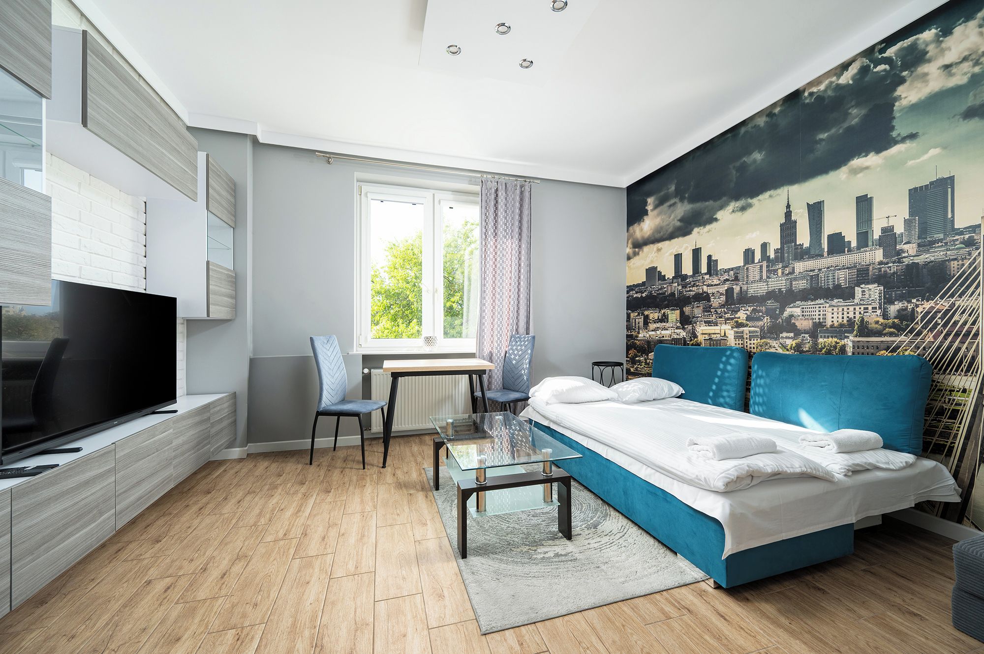 Ein modernes Schlafzimmer mit Stadtbild-Wandgemälde, einem großen Bett, einem Fernseher und einem Essbereich.