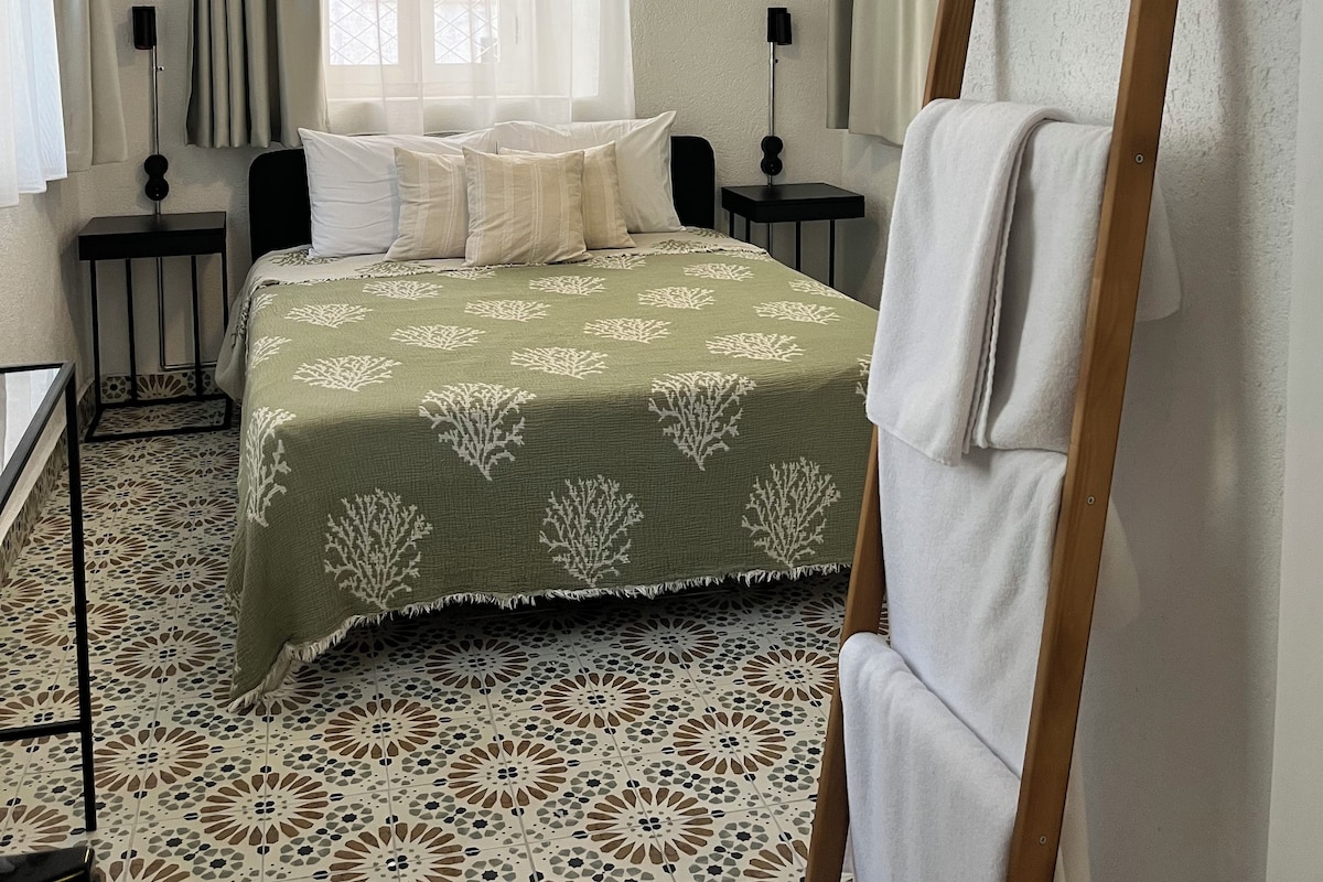 A room with a bed, two bedside tables, a standing lamp, a window with curtains, and a patterned floor.