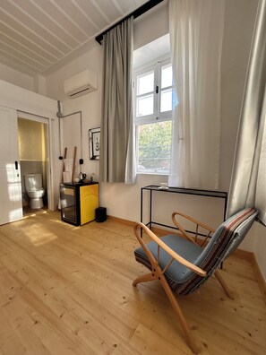 Una habitación con piso de madera, una silla, una mesita, una ventana con cortinas y una puerta que da a un baño.