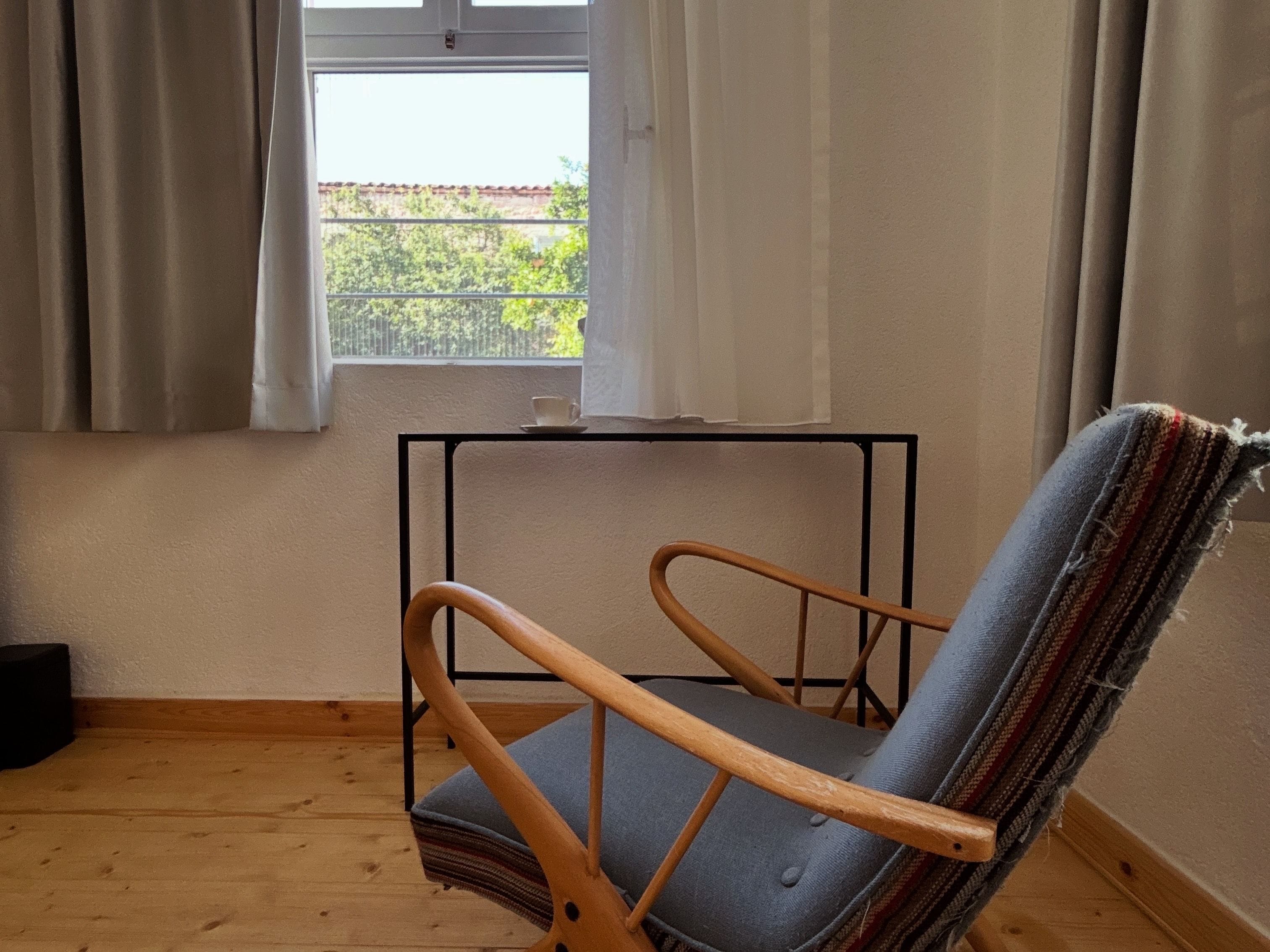 Une pièce avec un plancher en bois, une fenêtre avec des rideaux et une chaise dont l’assise est en osier tressé.