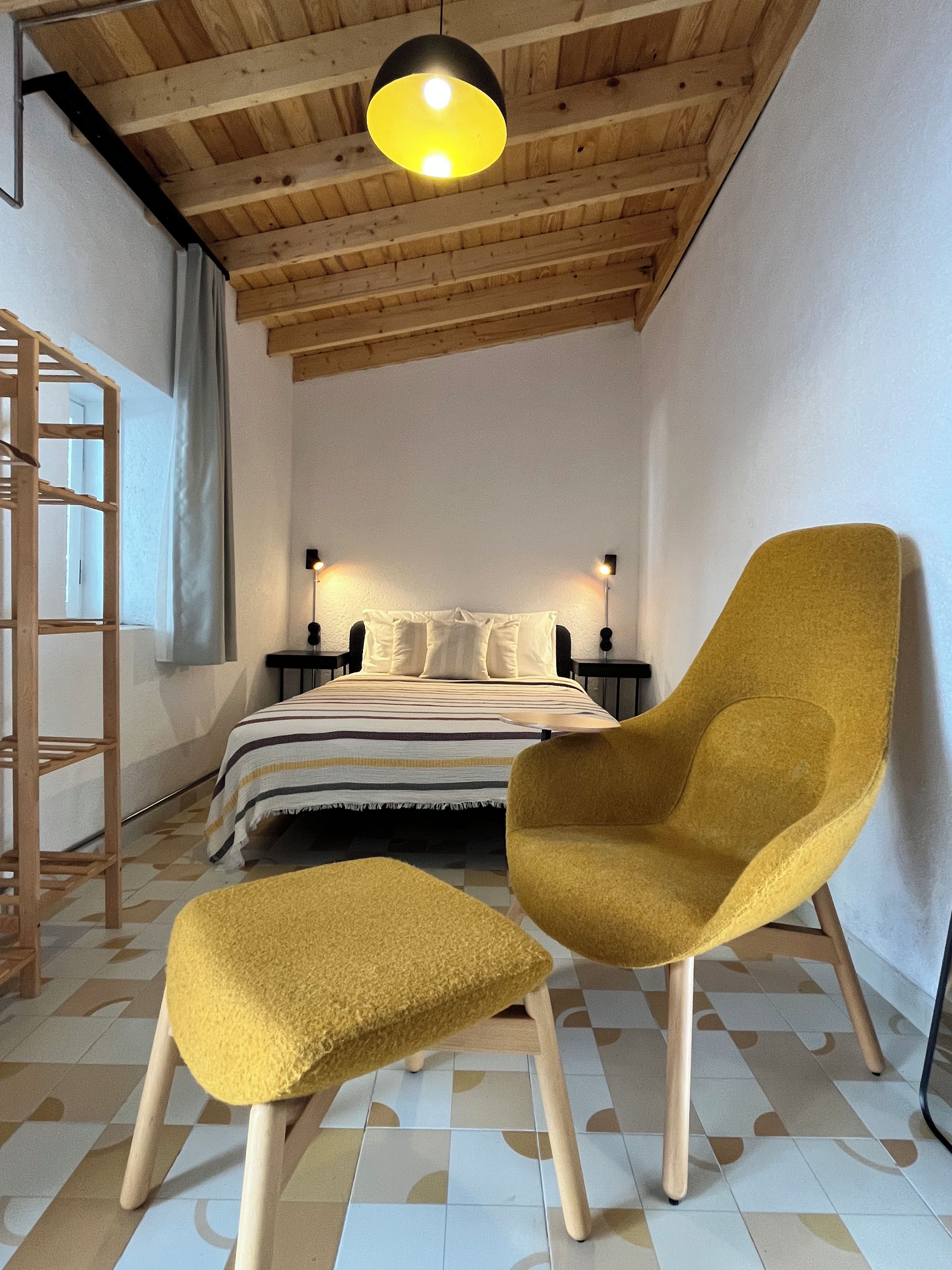Une chambre à coucher avec un lit, deux tables de chevet, une échelle en bois, un fauteuil jaune et un pouf assorti.