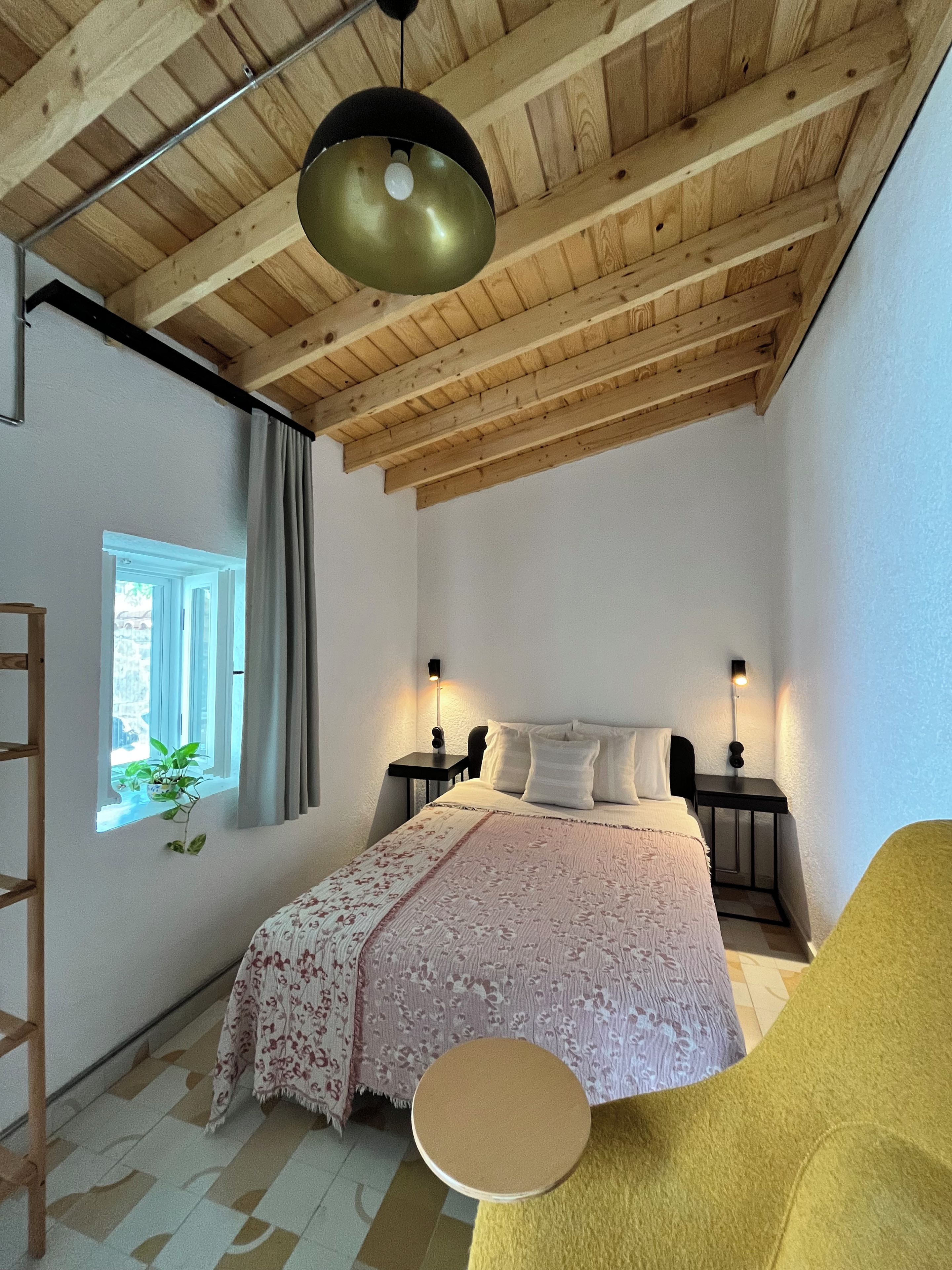 A bedroom with a wooden ceiling, a bed with a floral bedspread, two bedside tables with lamps, a window with curtains, and a yellow chair.