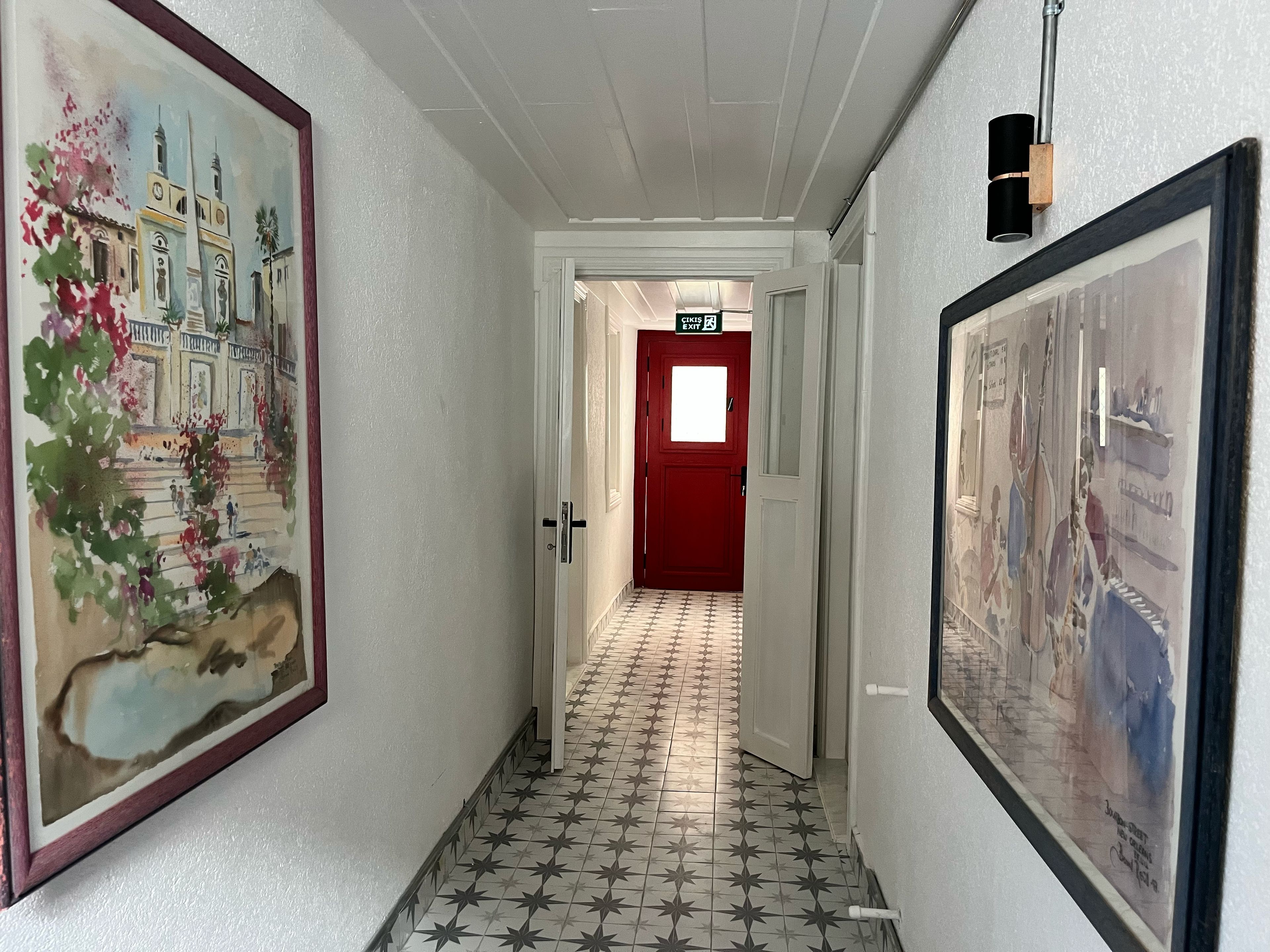 A hallway with a tiled floor, a red door, and framed artwork on the walls.