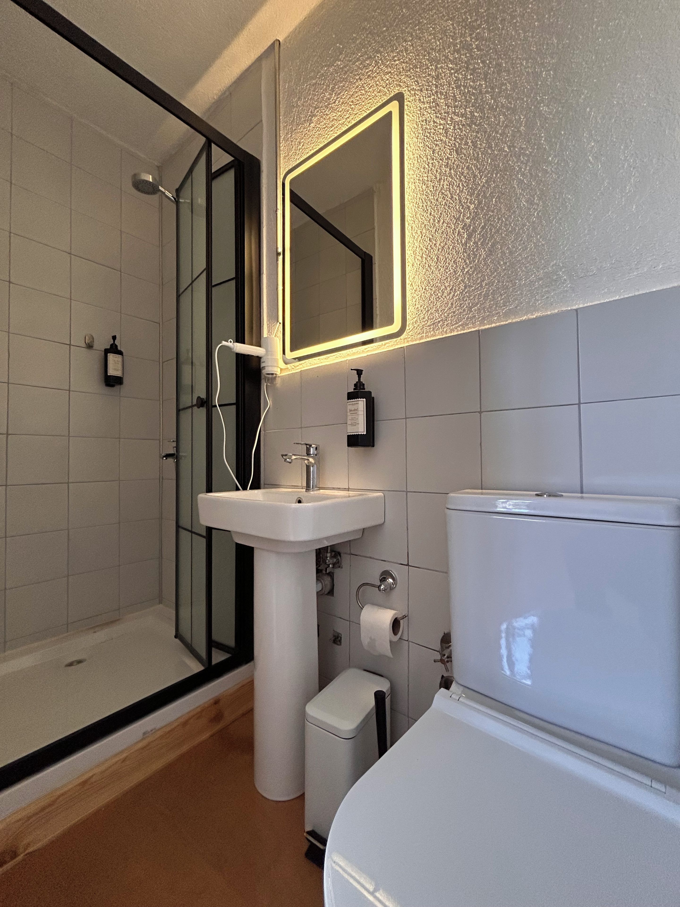 A modern bathroom with a glass shower enclosure, a pedestal sink, a toilet, and a mirror with a light.
