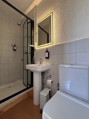 Un baño moderno con cabina de ducha de vidrio, lavamanos pedestal, inodoro y espejo con luz.