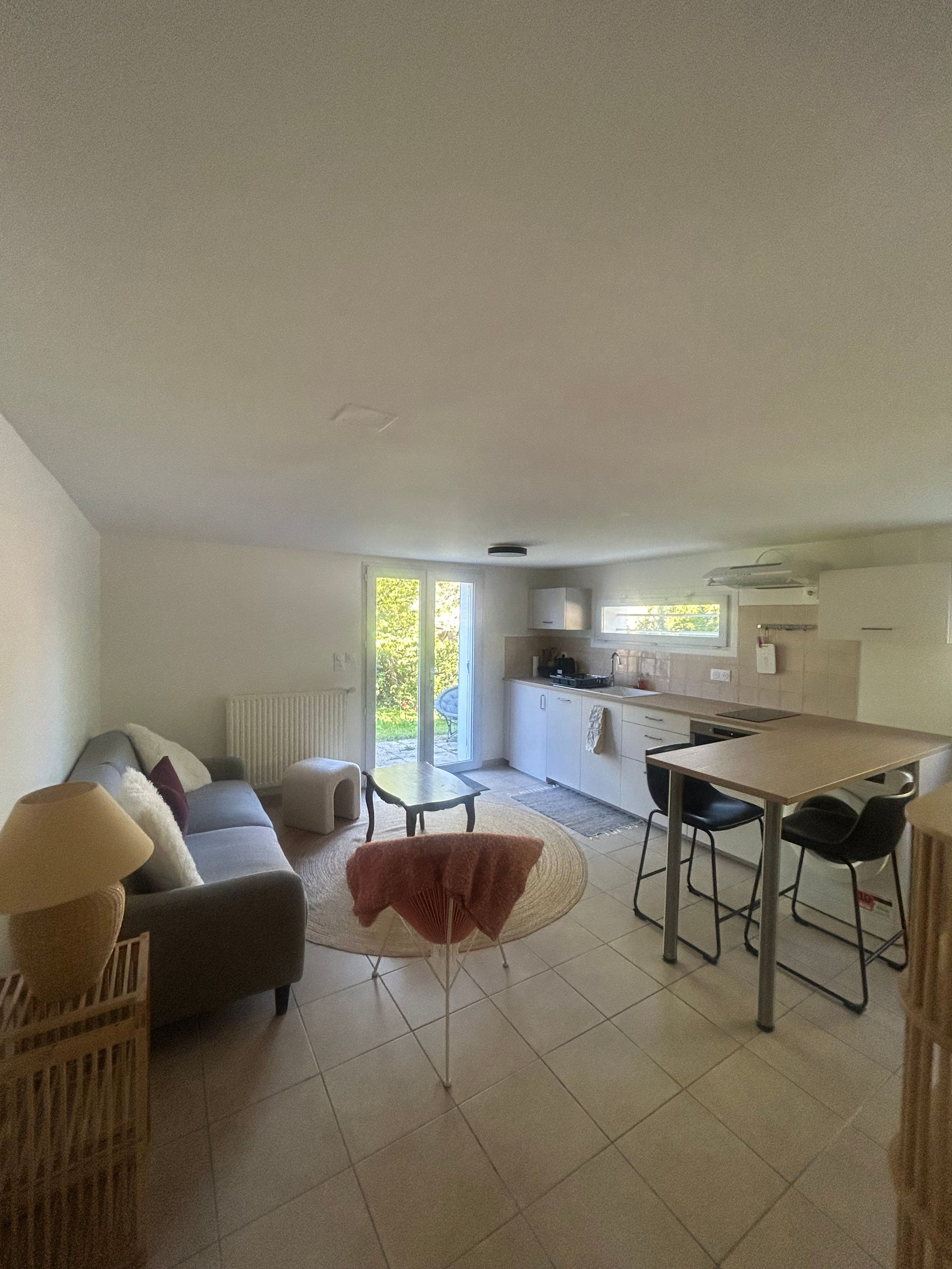 A modern living room with a sofa, a dining table, and chairs. There is a kitchen area with cabinets and a window.