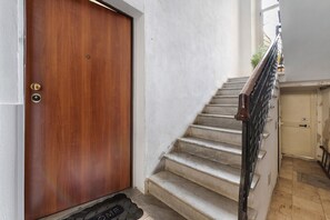 Une porte en bois avec une poignée en laiton et un trou de serrure, un escalier en marbre avec une rampe en métal noir, et un paillasson sur lequel est inscrit le mot « BIENVENUE ».