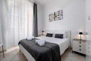 Ein modernes Hotelzimmer mit einem großen Bett, zwei Nachttischen mit Lampen, einem Fenster mit durchscheinenden Vorhängen und einer Wand mit drei gerahmten Kunstwerken.