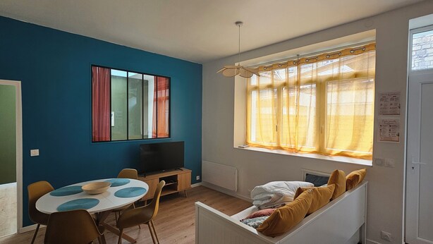 A room with a blue wall, a window with yellow curtains, a television on a stand, a dining table with chairs, and a bench with cushions. - Vrbo Property (Thury-Harcourt-le-Hom)