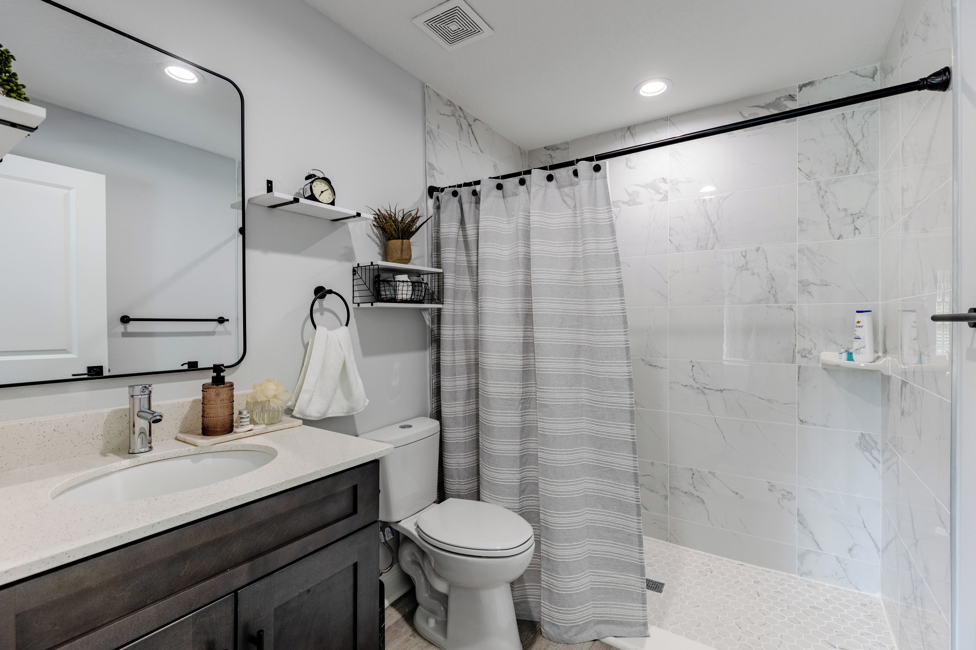 A modern bathroom with a white marble wall, a shower with a grey striped curtain, a white toilet, and a sink with a soap dispenser.