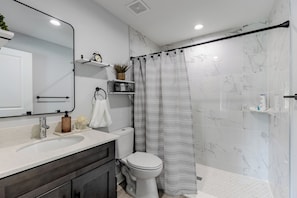 A modern bathroom with a white marble wall, a shower with a grey striped curtain, a white toilet, and a sink with a soap dispenser. - 6 mins to WEC Precious Mini house (Ocala)