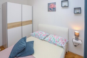 Un dormitorio con cama, almohadas, un armario, una mesita de noche con lámpara y una estantería de pared con adornos.