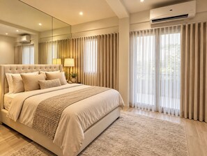 Une chambre à coucher moderne avec un grand lit, une table de chevet avec une lampe et une vue sur l’extérieur à travers des rideaux légers.