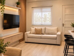 A modern living room with a flat-screen TV, a beige sofa, and a dining area with a table and chairs. - Modern 1BR Executive Suite in Taguig near BGC (Taguig)