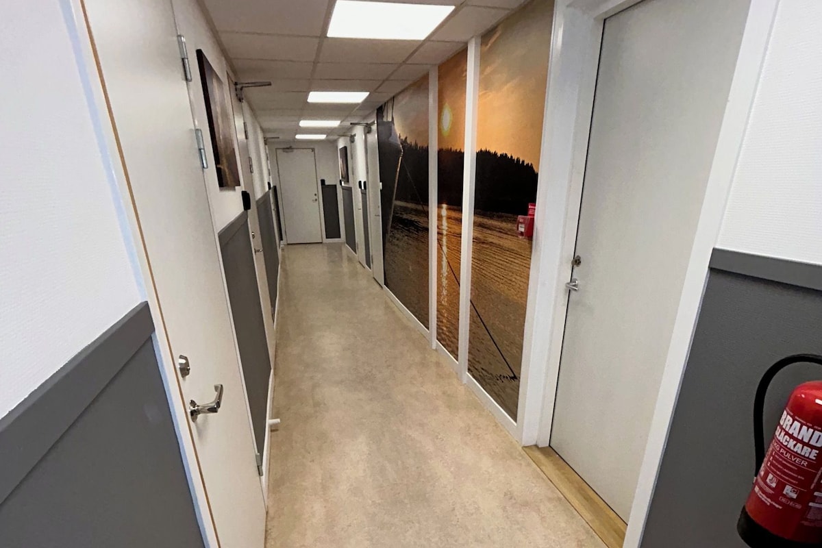Un pasillo con puertas a ambos lados, un extintor a la derecha y una señal de salida en el techo.