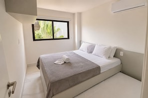 Un lit simple avec une couverture grise et des oreillers blancs, une fenêtre donnant sur un paysage verdoyant et un climatiseur fixé au mur.