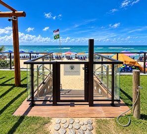 Une entrée située au bord de la plage, dotée d’une structure en verre, d’un drapeau et d’un panneau indiquant « Accès réservé ».