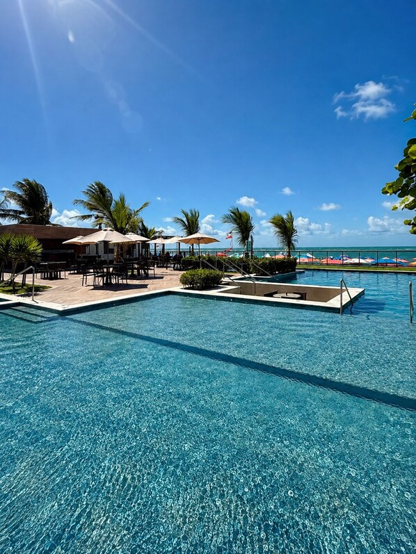 Une piscine d’un bleu éclatant, bordée de chaises longues et d’ombrelles, entourée de palmiers et offrant une vue sur l’océan.