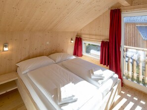 Ein Schlafzimmer einer Holzhütte mit einem Bett, zwei Kissen, einem Fenster mit Vorhängen und einer kleinen Ablagefläche.