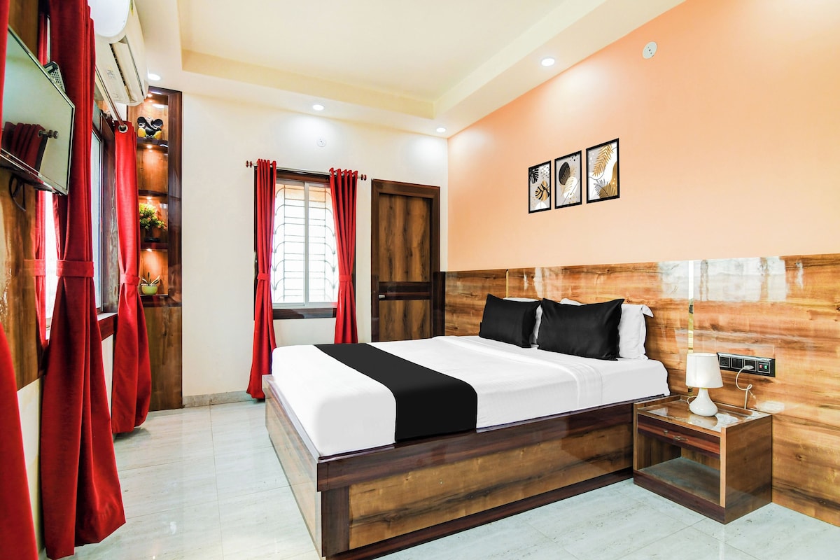 A hotel room with a wooden headboard, a bed with white and black bedding, a nightstand with a lamp, and a window with red curtains.