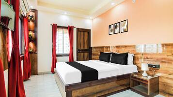 Habitación de hotel con cabecera de madera, cama con ropa de cama blanca y negra, mesita de noche con lámpara y ventana con cortinas rojas.