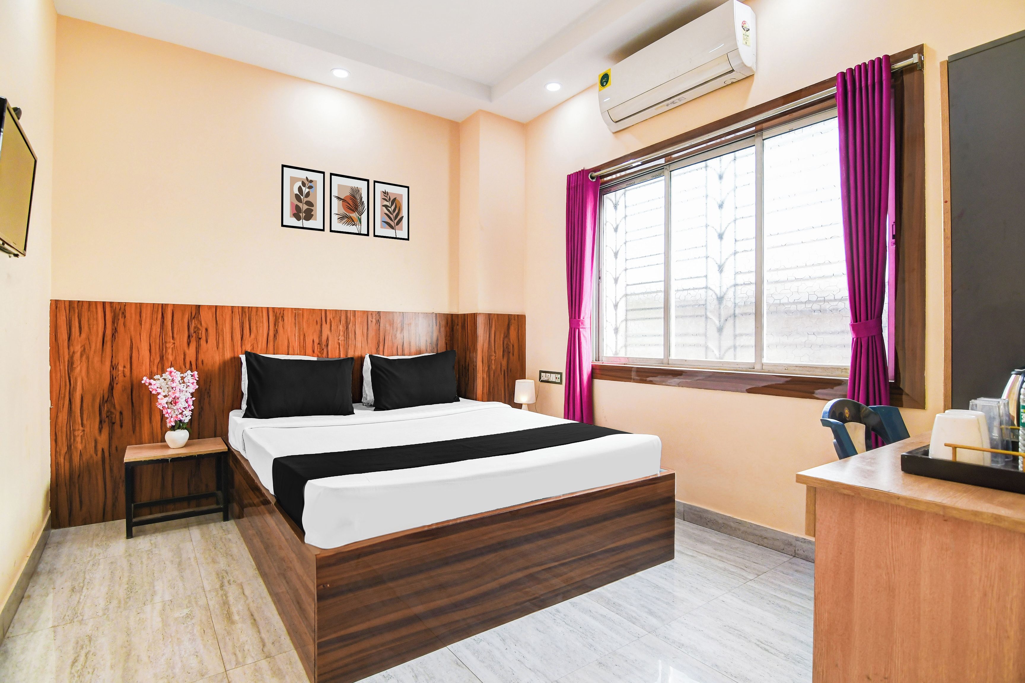 Habitación de hotel con cabecera de madera, cama con ropa de cama blanco y negro, mesita de noche con un jarrón de flores, escritorio con una silla y ventana con cortinas moradas.