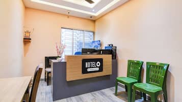 Un mostrador de recepción de hotel con un cartel que indica servicio de empresa, un ventilador de techo, una ventana con persianas y dos sillas verdes.