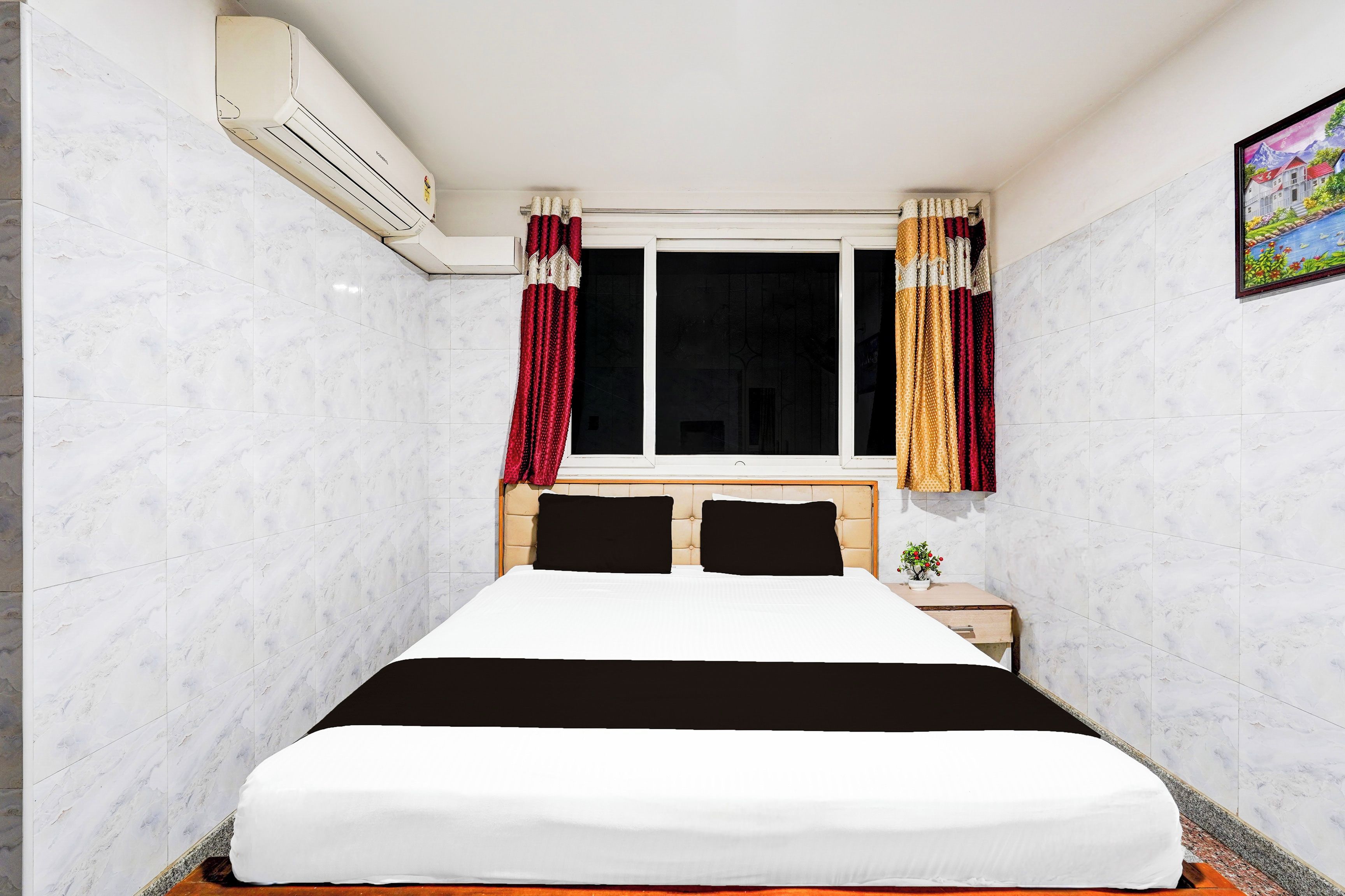 Una habitación de hotel con cama, ventana con cortinas, aire acondicionado y un cuadro en la pared.