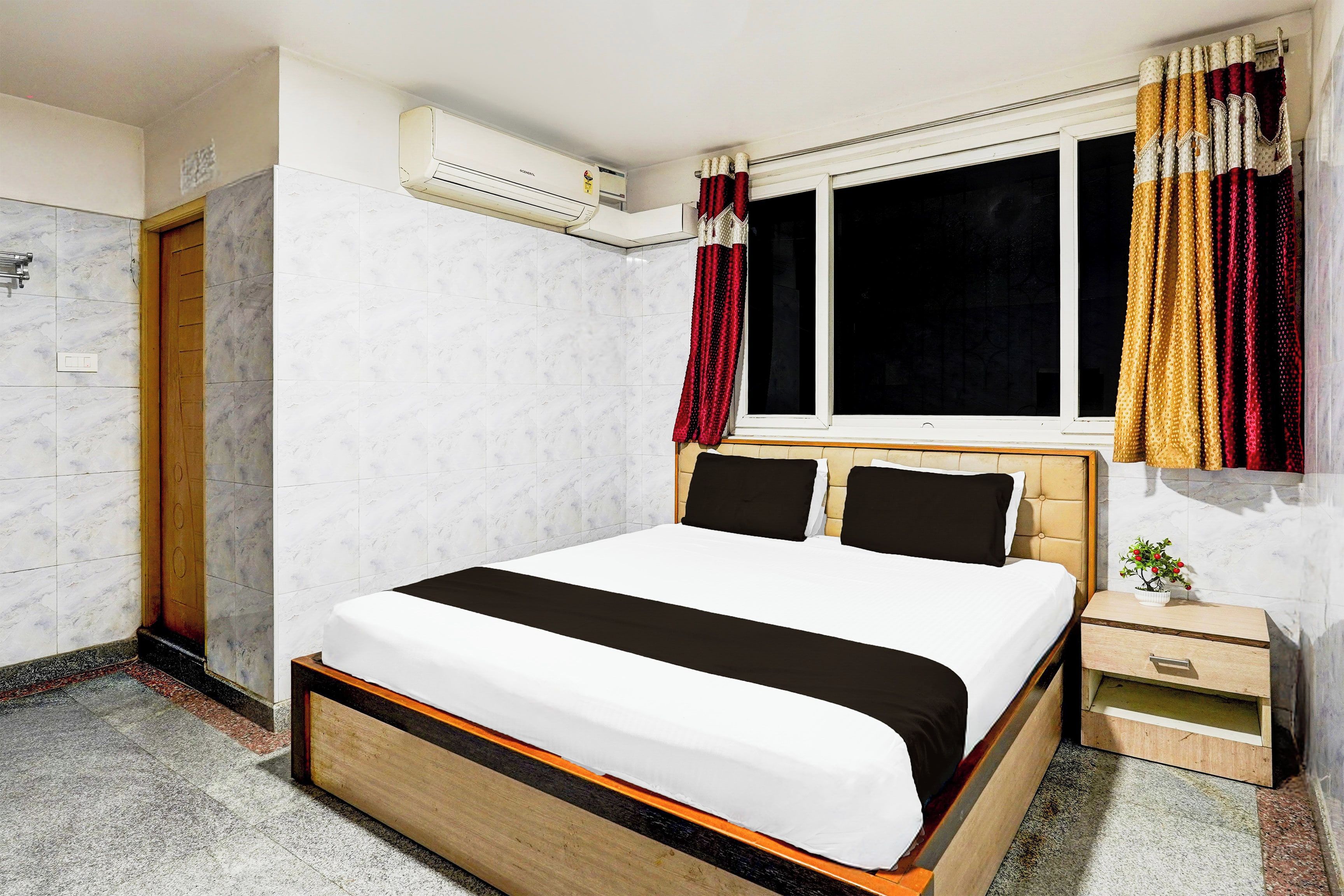 Habitación de hotel con cama, ventana con cortinas, unidad de aire acondicionado y una mesita de noche con una planta.