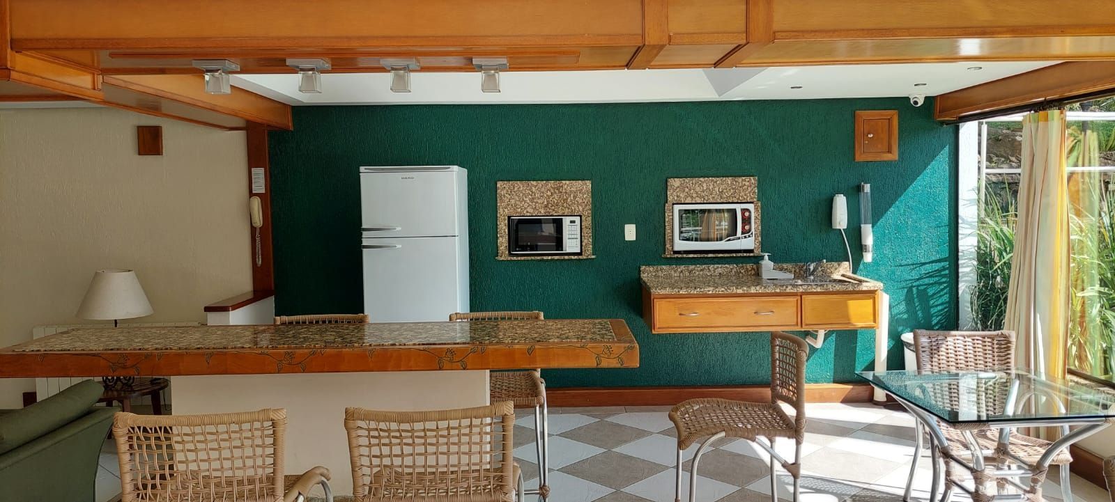 A compact kitchen with a refrigerator, microwave, and a small countertop. There are wicker chairs and a glass table.