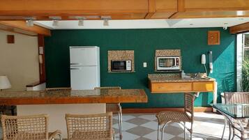 A compact kitchen with a refrigerator, microwave, and a small countertop. There are wicker chairs and a glass table.