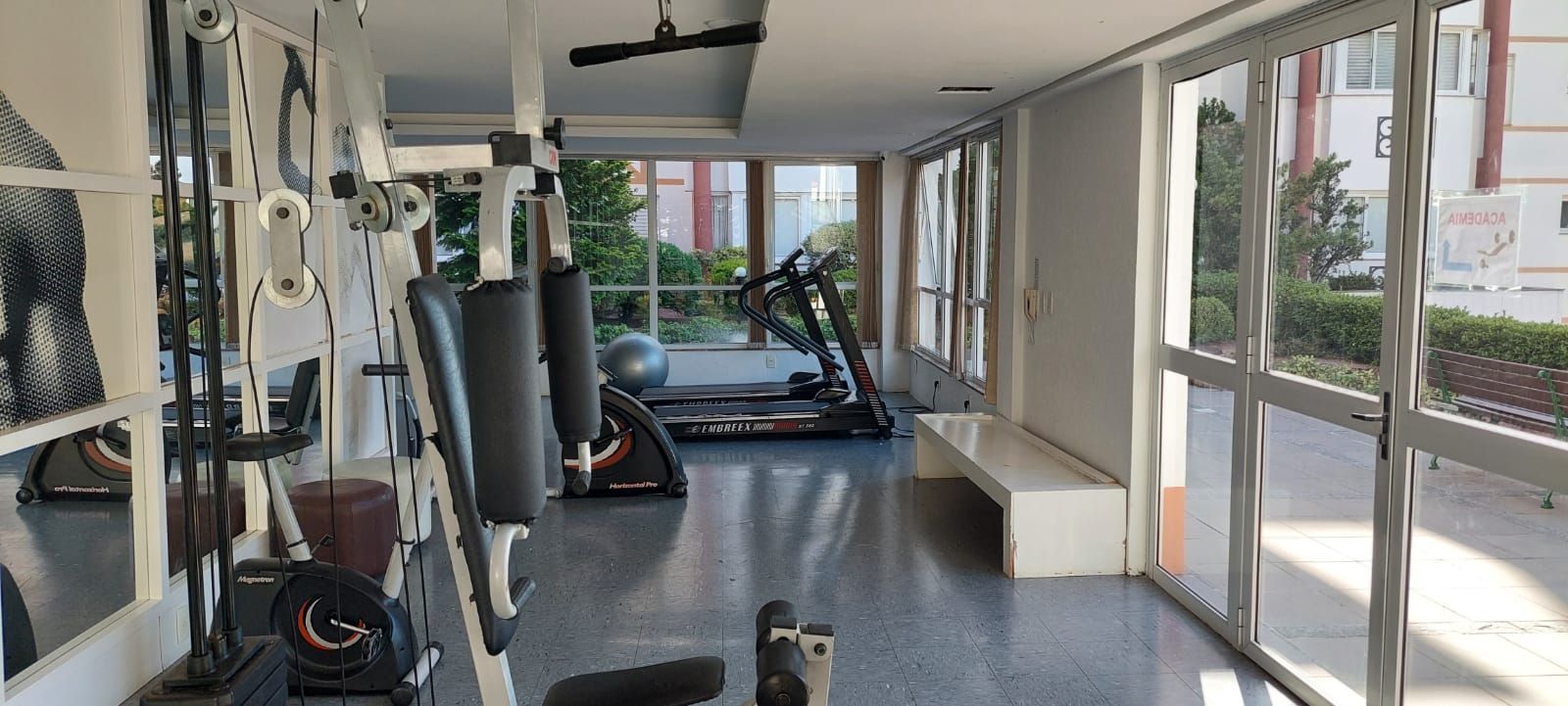 Uma academia com equipamentos de exercício, esteira e amplas janelas.