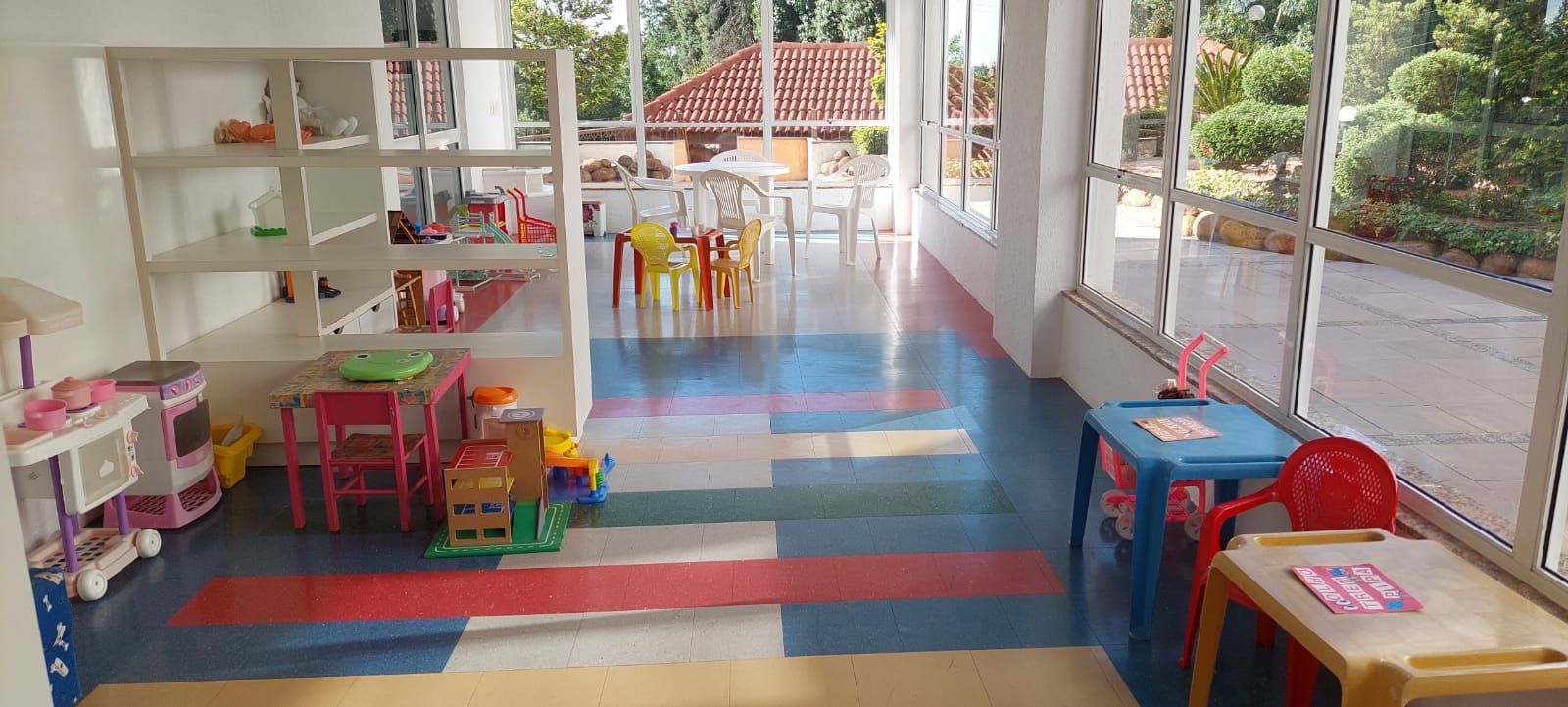 A children's play area with colorful flooring, shelves with toys, and a view of the outdoors through large windows.
