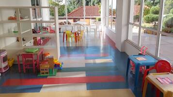 A children's play area with colorful flooring, shelves with toys, and a view of the outdoors through large windows.