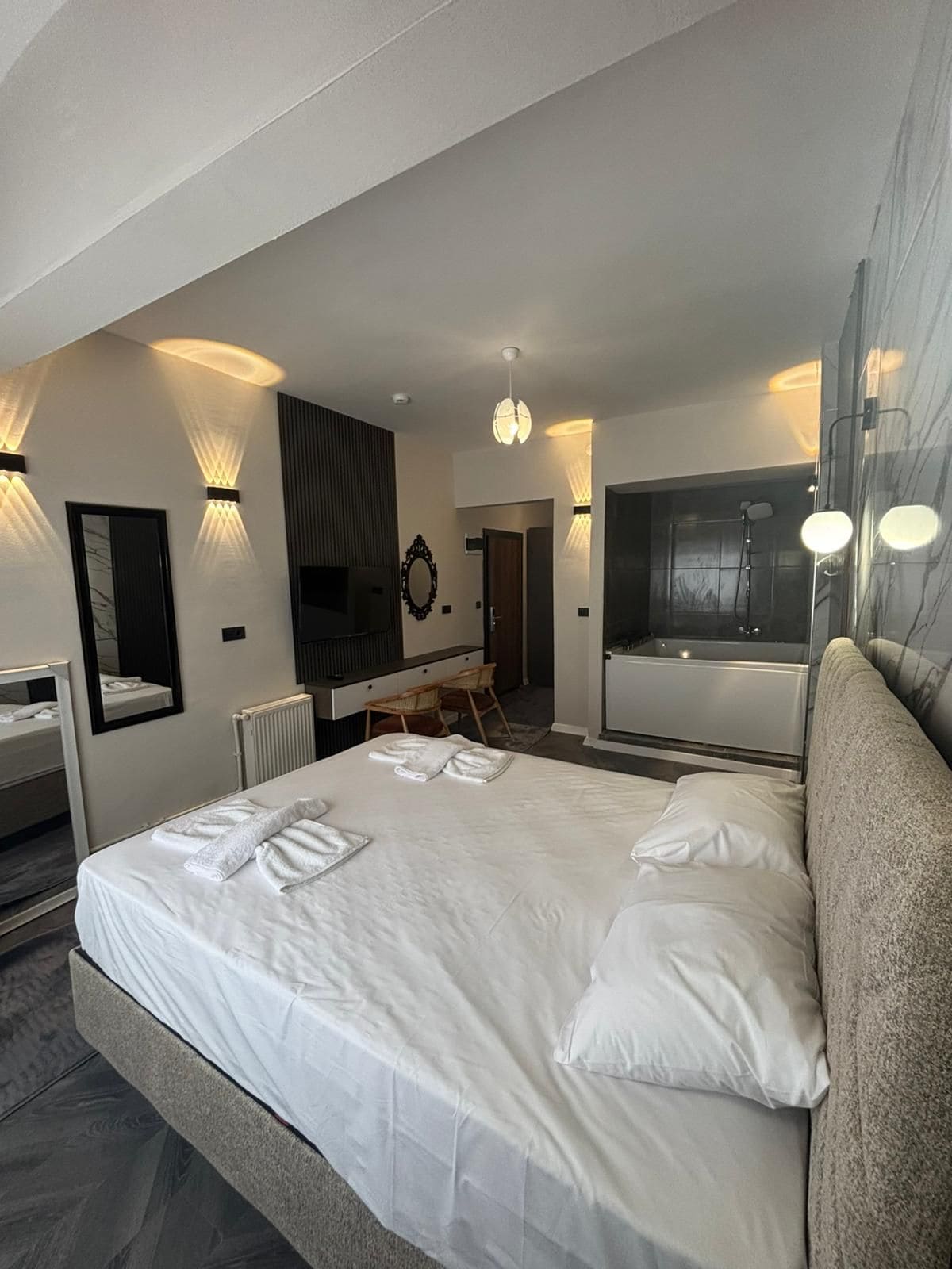 Une chambre d’hôtel moderne dotée d’un grand lit, d’un coin salon avec une table et des chaises, d’un miroir, ainsi que d’une salle de bain avec douche et toilettes.