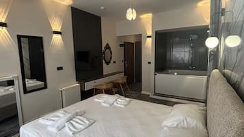 A modern hotel room with a large bed, a sitting area with a table and chairs, a mirror, and a bathroom with a shower and a toilet.