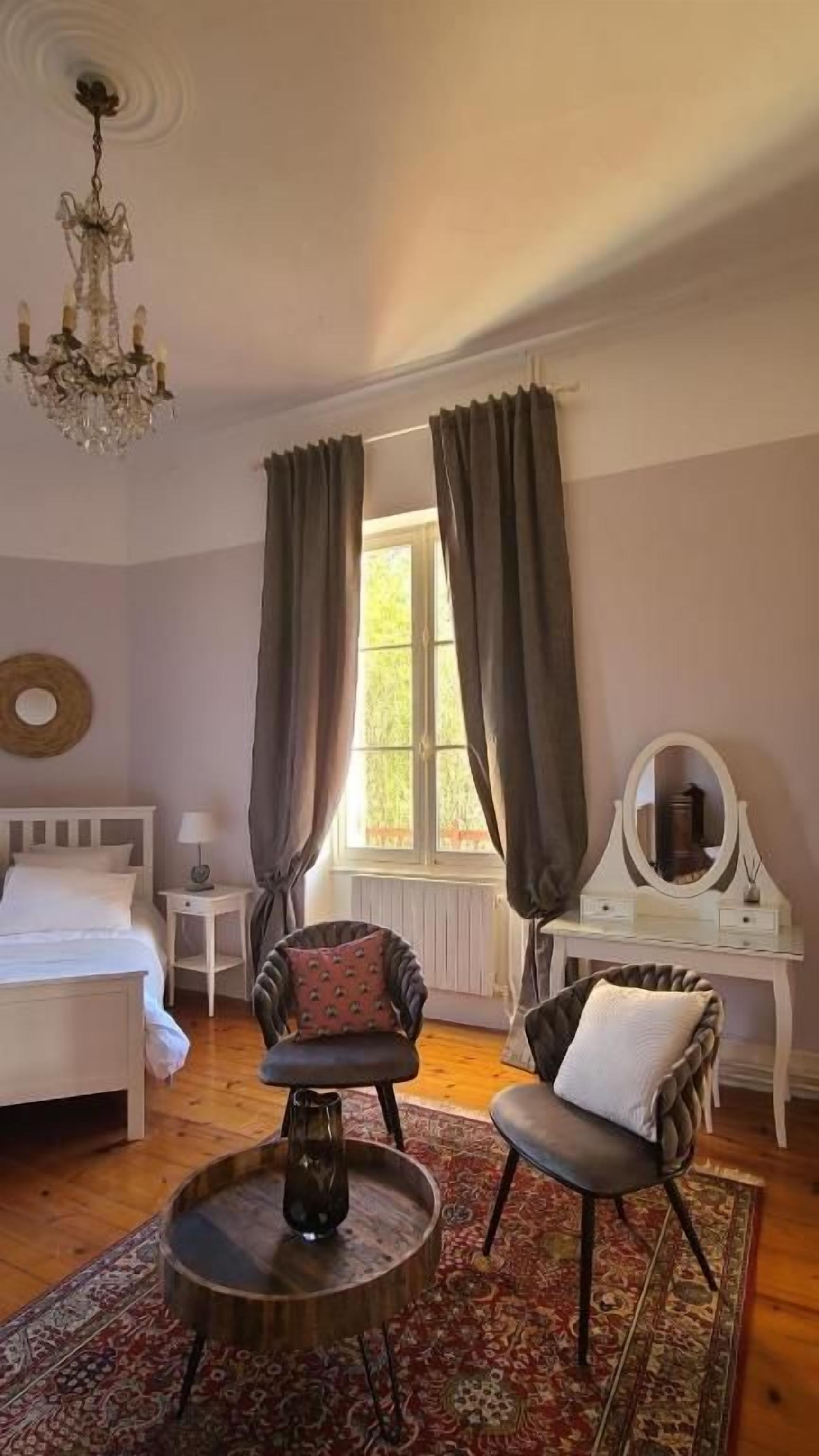 Ein Schlafzimmer mit einem Bett, zwei Sessel, einem runden Tisch, einer Kommode, einem Spiegel und einer Kronleuchter.