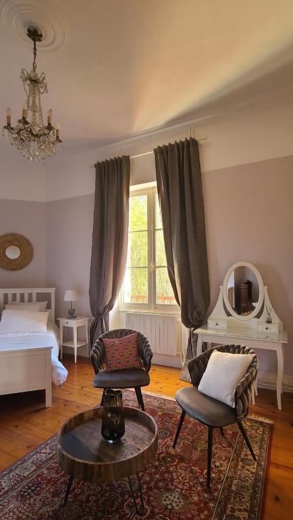 A bedroom with a bed, two chairs, a round table, a dresser, a mirror, and a chandelier.